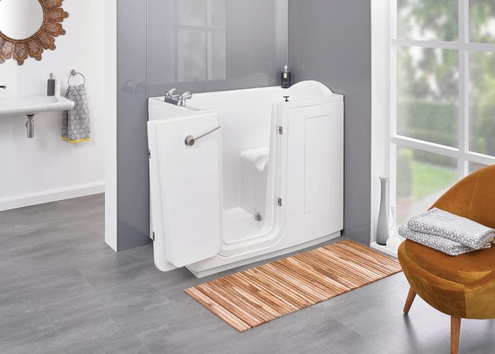 Disabled shower bath, contoured seat and wide door of the Palermo Walk-in Bath (Tennessee Midi Access Walk in Baths). Life Quality Bathing. Modern grey wall and flooring, Door window view of garden and brown leather chair next to the bath with towels folded on the edge.