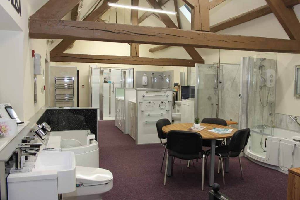 Bathroom showroom featuring accessible fixtures including bathtubs, sinks, showers, and toilets, with central seating area and exposed wooden beams
