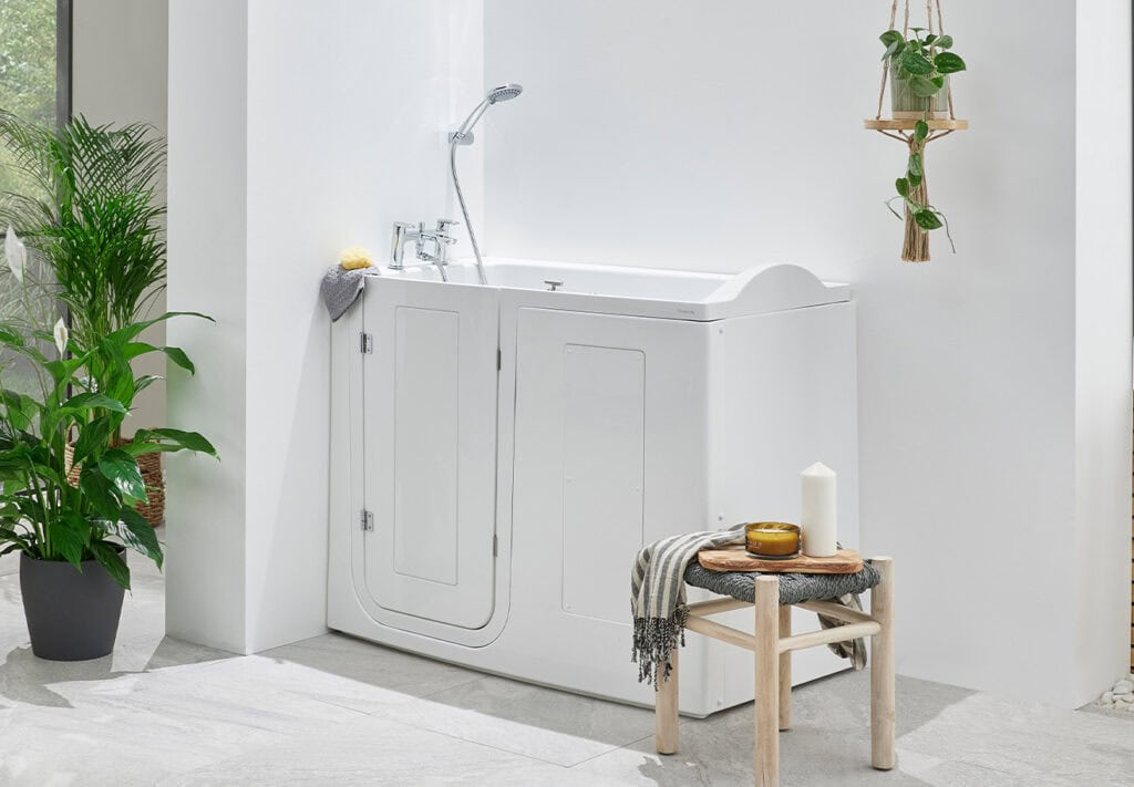 Palermo walk-in bath (Tennessee) with side door. Hand held shower head wall mounted one feet above bath. White decor, wooden stool with towel and candles.