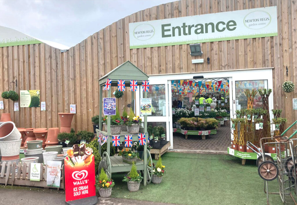 Garden Centre entrance. Small plant and pottery displayed before the corrugated wooden wall, with sliding open doors under large Entrance sign.